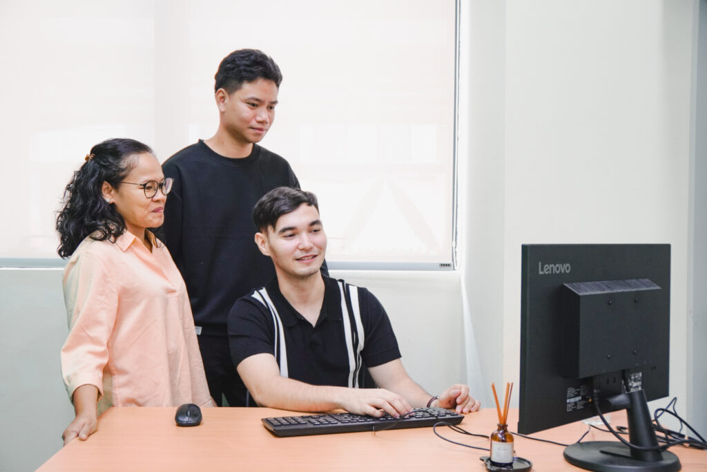 Three VizServe professionals in business casual attire collaborating in an office; one is seated using a laptop while two stand beside them looking at the screen.