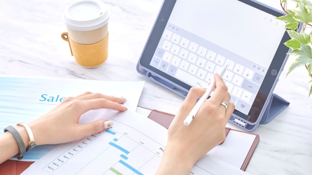 Person using a tablet and stylus to review sales documents and a Gantt chart in a modern workspace.